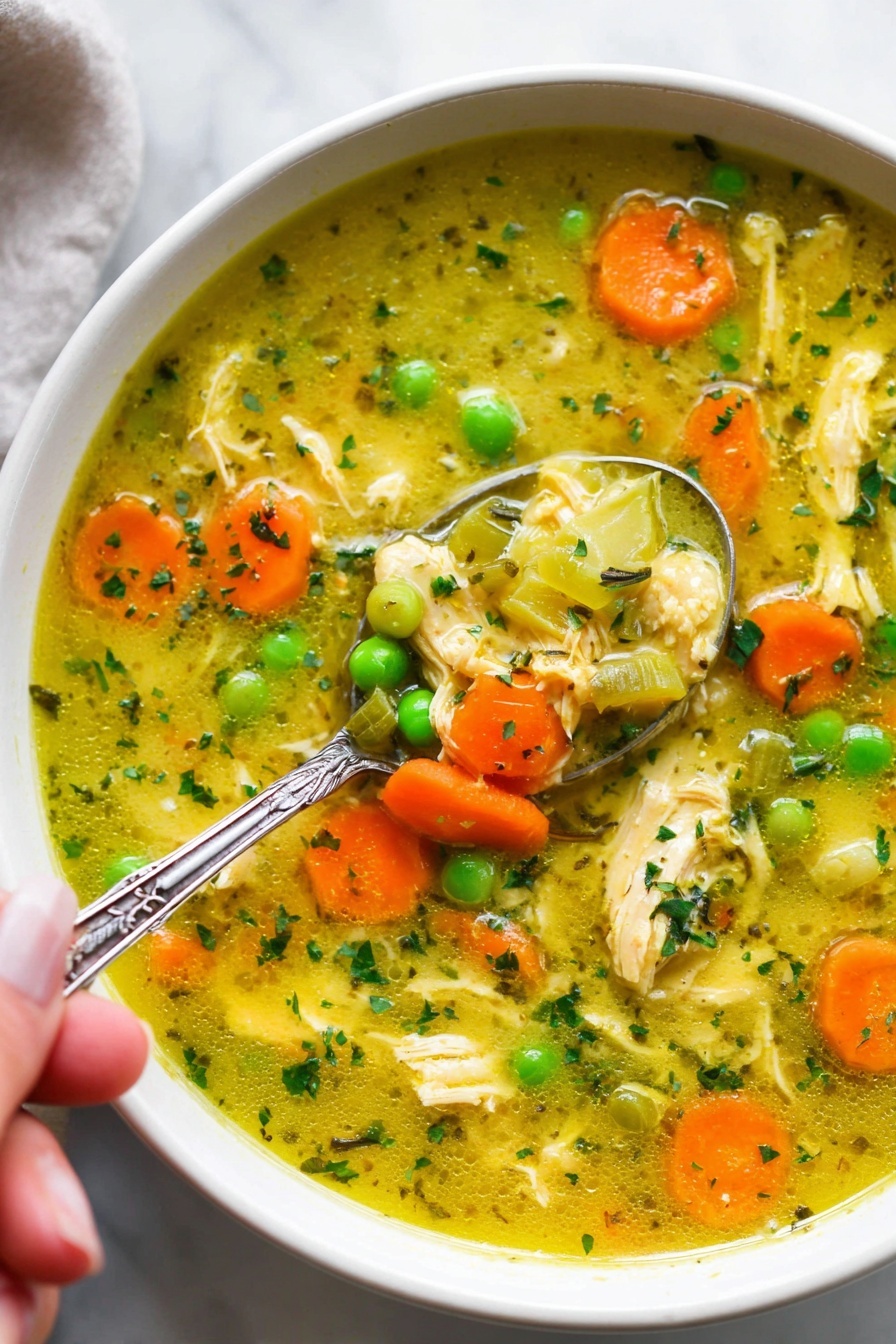 Turmeric Chicken Soup, healthy turmeric chicken soup, nourishing chicken soup with turmeric, anti-inflammatory chicken soup, comforting chicken soup recipe - The image shows a white bowl filled with a colorful chicken soup. The soup has a yellowish broth with small pieces of shredded chicken scattered throughout. Bright orange carrot slices and vibrant green peas float on the surface along with translucent chunks of celery. Small green herb bits are sprinkled all over, adding texture and color. A silver spoon with detailing on the handle is partially submerged in the soup, held by a woman's hand, scooping some of the ingredients. The bowl sits on a white marbled surface, giving a clean and fresh look. photo taken with an iphone --ar 2:3 --v 7