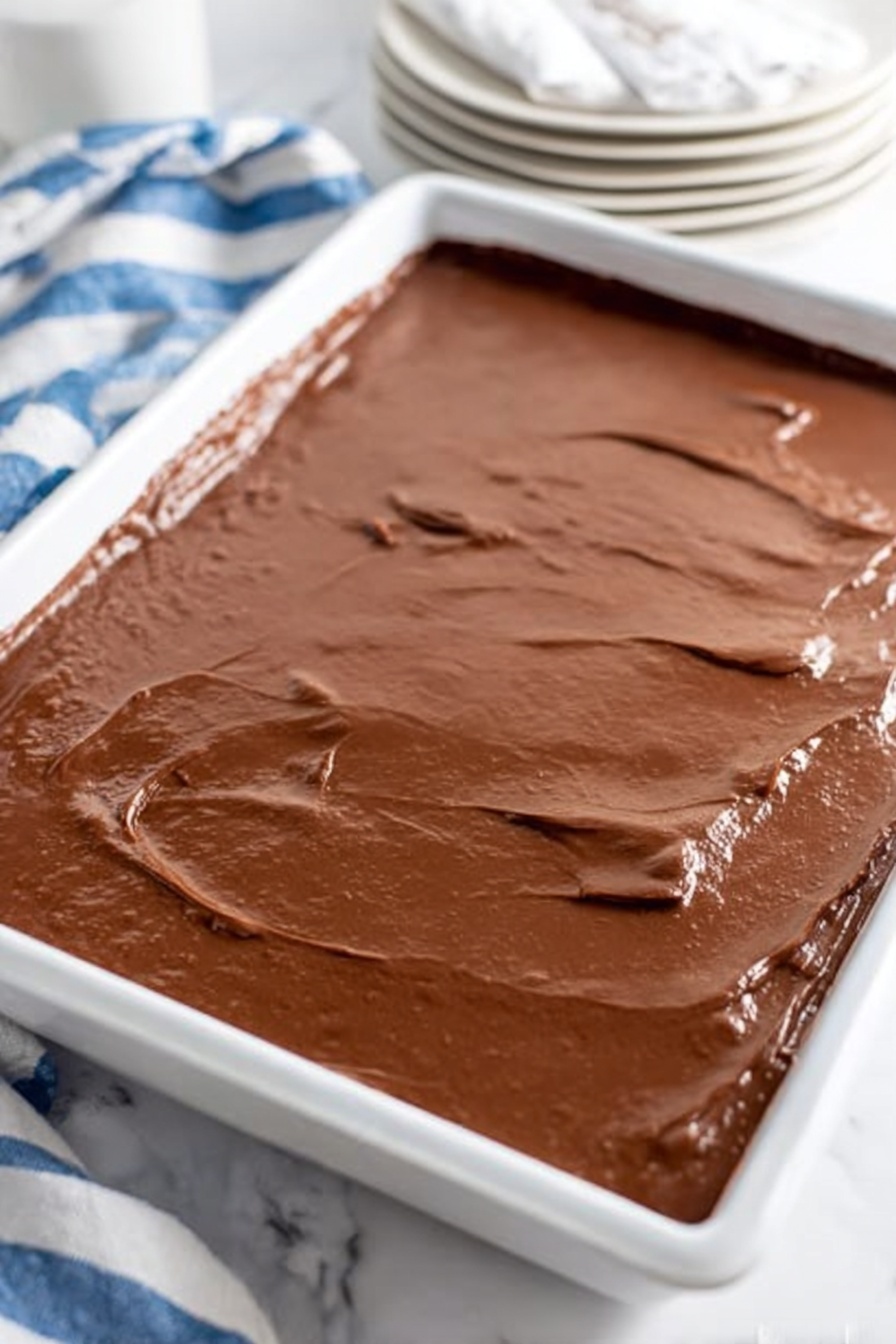 Texas Sheet Cake with Rich Cocoa Frosting, best Texas sheet cake, simple chocolate sheet cake recipe, easy chocolate cake for parties, moist chocolate sheet cake - A large white tray filled with a smooth, thick layer of shiny chocolate batter, spread evenly with slight ridges and soft peaks across the entire surface. The background includes a white marbled texture with a stack of white plates and white paper napkins on the top right, and a striped cloth in blue and white behind them. The chocolate layer looks rich and moist with a soft texture, covering the whole tray in one even layer. Photo taken with an iphone --ar 2:3 --v 7