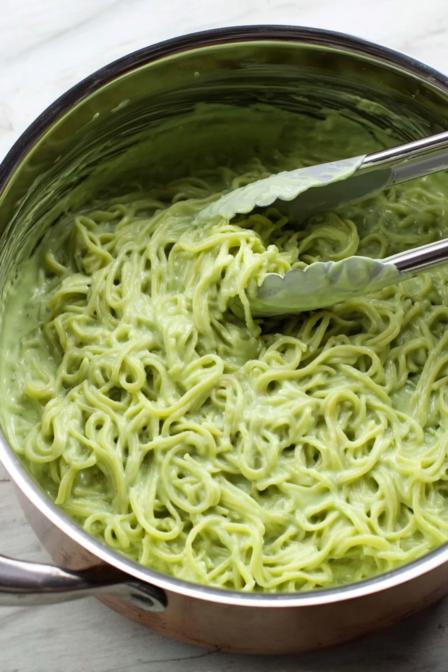Vegan Cilantro Lime Noodles, vegan noodle recipes, healthy vegan pasta, easy vegan dinner ideas, fresh plant-based meals - Inside a shiny silver pot placed on a white marbled surface, there is a mass of noodles covered in a thick, light green sauce. The sauce is creamy and clings to the noodles, giving them a smooth texture. A silver tong is partially dipped into the noodles on the right side of the pot, with a bit of sauce stuck on the tong. The noodles have a slightly twisted shape and fill most of the pot inside. Photo taken with an iphone --ar 2:3 --v 7