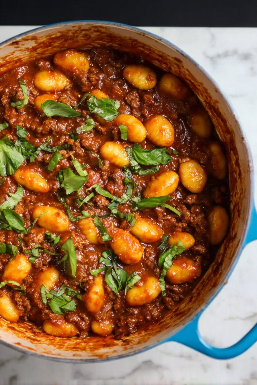 Gnocchi Bolognese, hearty pasta dish, baked gnocchi with meat sauce, comforting Italian dinner, easy gnocchi recipes - The image shows a blue pot filled with a rich brown meat sauce mixed with soft, yellow-orange gnocchi. The top layer is scattered with pieces of fresh green herbs, adding a bright contrast to the warm colors of the sauce and gnocchi. The pot's handle is visible on the right side, resting on a white marbled surface. The texture of the gnocchi looks smooth and tender, while the meat sauce appears thick and hearty, covering all pieces evenly. Photo taken with an iphone --ar 2:3 --v 7