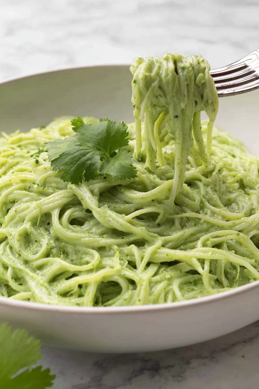 Vegan Cilantro Lime Noodles, vegan noodle recipes, healthy vegan pasta, easy vegan dinner ideas, fresh plant-based meals - This image shows a white bowl filled with creamy green noodles, with about two layers of noodles visible, all coated evenly in a thick, smooth green sauce. On top of the noodles, there is a single thin slice of lime placed near the center, with bits of black pepper sprinkled across the noodles. Next to the lime slice, a small bunch of fresh green cilantro leaves rests gently. A silver fork is placed on the right edge of the bowl, partly inserted into the noodles. The bowl sits on a white marbled surface. Photo taken with an iphone --ar 2:3 --v 7