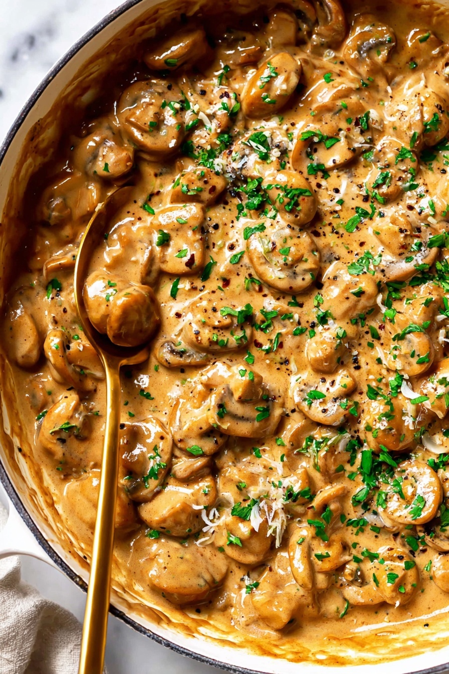 French Onion Pasta with Melted Gruyere, caramelized onion pasta, cheesy pasta recipes, savory onion pasta dish, easy gourmet pasta - The image shows a close-up of a white pan filled with creamy mushroom sauce. The sauce has many small round and sliced mushrooms in a thick, light brown sauce with visible black pepper and small bits of herbs. The top layer is sprinkled with fresh green chopped parsley. A brass-colored spoon is partially dipped in the sauce toward the left side of the pan. The pan is placed on a white marbled surface. Photo taken with an iphone --ar 2:3 --v 7