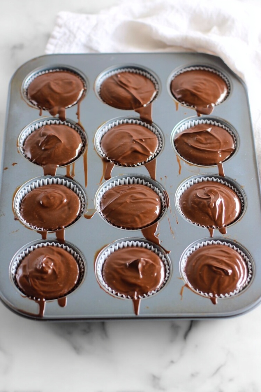 No Bake Peanut Butter Oat Cups, no bake energy bites, healthy no bake snacks, quick breakfast treats, peanut butter oat energy bites - A gray muffin tray with twelve round cups, each filled with a smooth, glossy layer of melted chocolate that slightly spills over the edges. The chocolate has a shiny, swirled texture on top, and thin chocolate drips and streaks connect some of the cups and run down the tray’s sides. The tray rests on a white marbled surface with a soft white cloth partially visible at the top right corner. Photo taken with an iphone --ar 2:3 --v 7