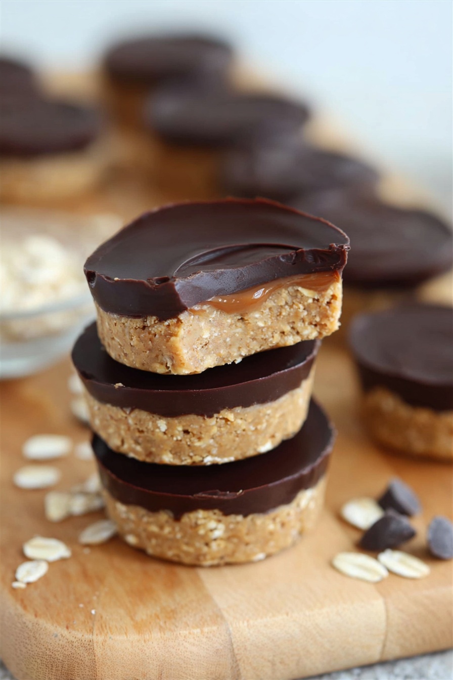 No Bake Peanut Butter Oat Cups, no bake energy bites, healthy no bake snacks, quick breakfast treats, peanut butter oat energy bites - A close-up of a woman's hand holding a small round dessert with two layers, the bottom layer is a light tan, crumbly oat mixture, and the top layer is smooth, dark chocolate spread evenly. In the background, several more of these two-layer treats are stacked and scattered on a wooden board set on a white marbled surface, with some oats sprinkled nearby. The overall scene is clean and bright with soft lighting, highlighting the texture contrast between the oat base and the chocolate top. Photo taken with an iphone --ar 2:3 --v 7