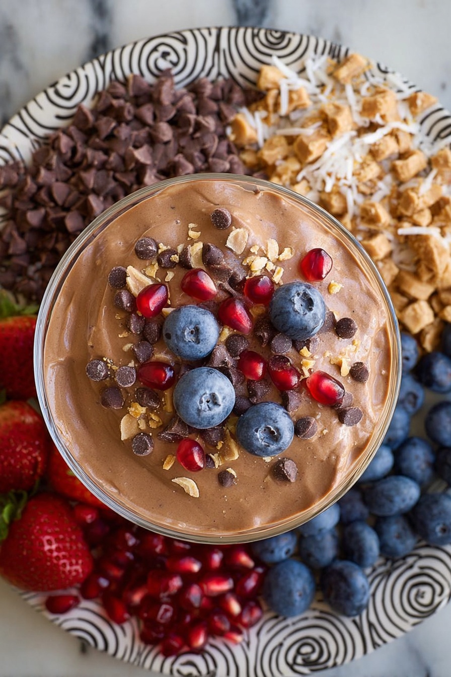 Chocolate Protein Fluff, healthy chocolate dessert, high-protein snacks, quick protein dessert, fluffy chocolate treat - A large round white plate with black spiral and zigzag patterns holds eight sections of colorful food arranged around a central glass bowl filled with smooth light brown chocolate mousse. Starting from the top left going clockwise, there are bright red whole strawberries with green tops, small dark brown chocolate chips, off-white shredded coconut, chopped pieces of chocolate peanut butter cups, bright red pomegranate seeds, dark blue blueberries, beige crunchy granola clusters, and then back to the strawberries. The plate is placed on a white marbled surface. Photo taken with an iphone --ar 2:3 --v 7