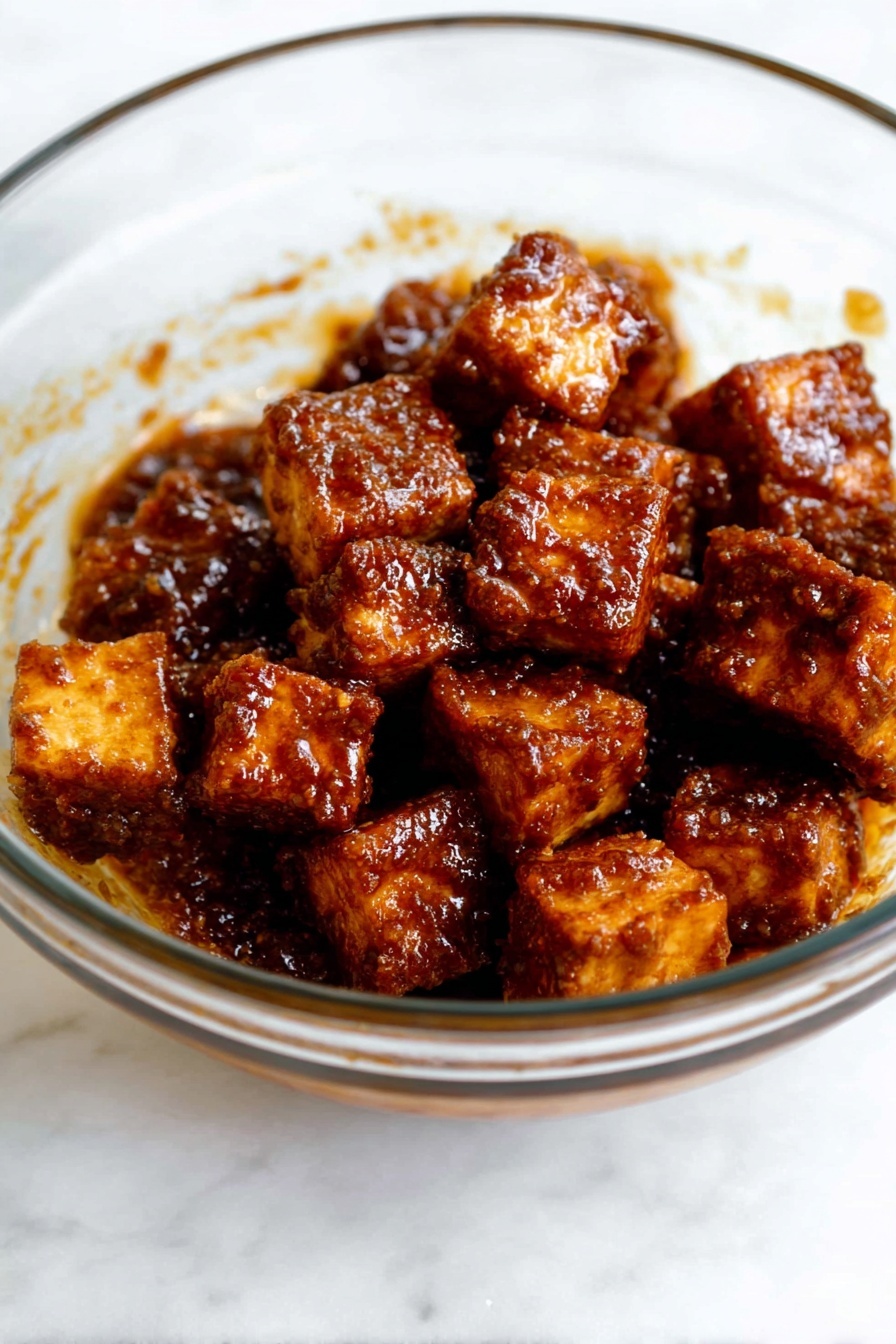 Crispy Gochujang Korean Tofu, Korean tofu with gochujang, spicy Korean tofu, vegan crispy tofu, easy Korean tofu recipe - The image shows a clear glass bowl filled with several small cubes of fried tofu. Each tofu piece is coated in a dark brown, shiny sauce with a textured surface that looks sticky and flavorful. The tofu pieces are piled together in the bowl, some slightly stacked on top of others. The smooth surface of the glass bowl lets you see the sauce clinging to the sides. The bowl is placed on a white marbled surface. photo taken with an iphone --ar 2:3 --v 7