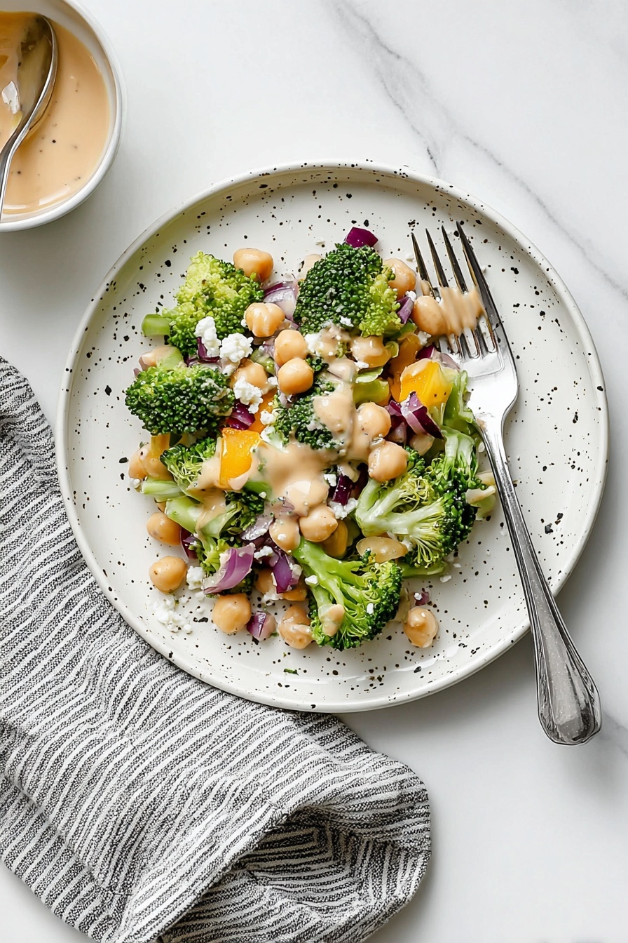 Broccoli and Chickpea Salad with Lemon Tahini, healthy vegan salad recipes, easy veggie chickpea salad, citrus tahini salad, quick plant-based recipes - A white bowl filled with a colorful chopped salad showing three clear layers: a base layer of small broccoli florets and chunks of celery creating a dense green texture, a middle layer with light-colored chickpeas and bright orange bell pepper pieces scattered evenly, and a top layer with small cubes of purple onion and white crumbled cheese spread across, topped lightly with thin almond slices and a creamy tan dressing drizzled over. Around the bowl are a gray and white striped cloth, a white marbled surface, a spoon in a small jar of creamy dressing, a small gray dish with salt and pepper, and a fork and knife nearby, photo taken with an iphone --ar 2:3 --v 7