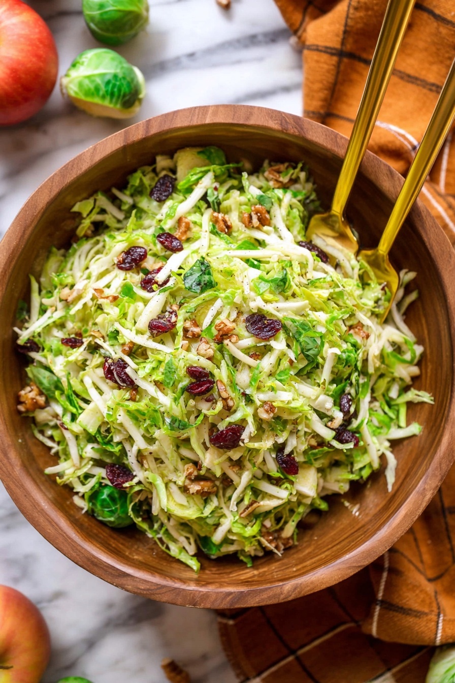 Brussels Sprouts Kale Apple Salad with Bacon, healthy Brussels sprouts salad, easiest kale apple salad, savory bacon apple salad, crunchy Brussels sprouts and kale salad - The image shows a wooden bowl full of a shredded green salad mixed with small bits of white cheese and dark red pieces, likely dried cranberries or bacon bits, with a texture of thin green leaves and stems. Two gold spoons rest in the bowl, positioned diagonally and partially buried in the salad. The bowl sits on a beige cloth over a white marbled surface, and in the background, bright orange small pumpkins and a few green Brussels sprouts are scattered on the same white marbled surface. The scene has a warm, natural feel with soft lighting. photo taken with an iphone --ar 2:3 --v 7