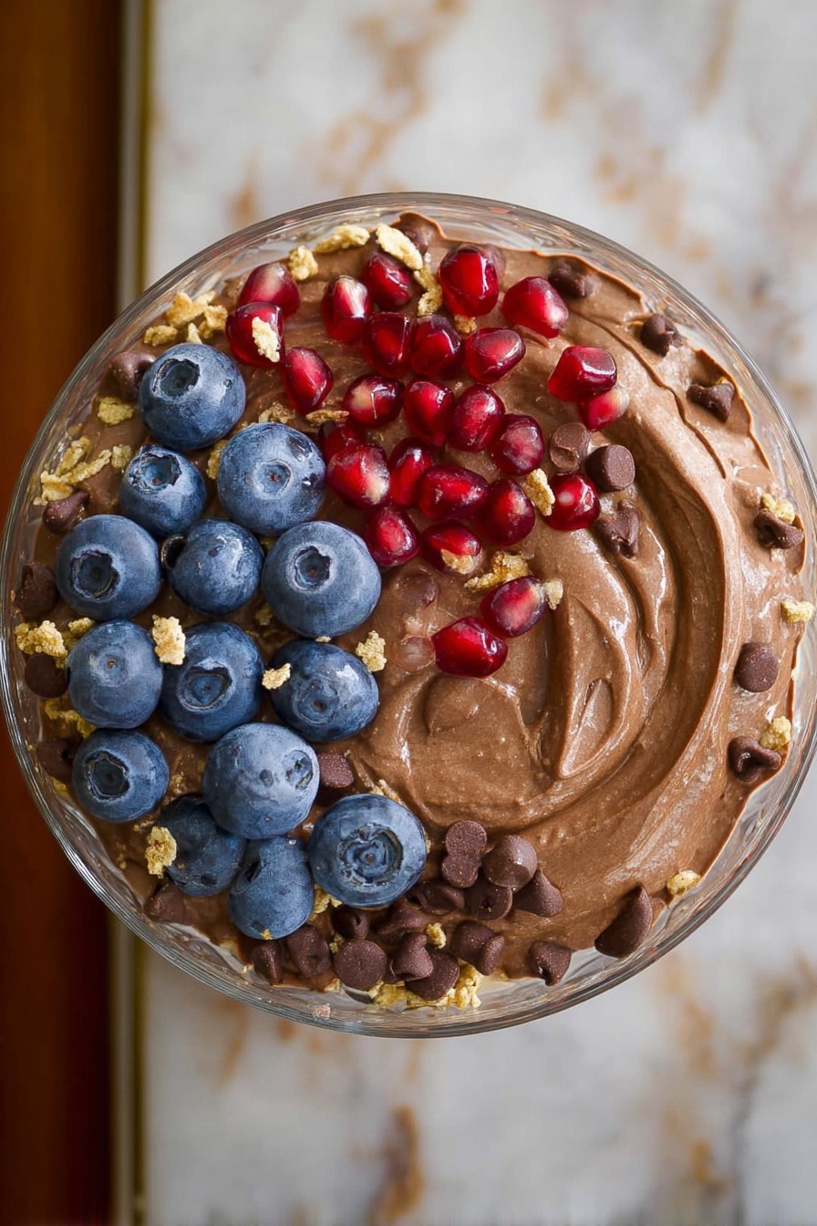 Chocolate Protein Fluff, healthy chocolate dessert, high-protein snacks, quick protein dessert, fluffy chocolate treat - A clear glass bowl in the center holds a creamy, smooth light brown pudding topped with a few scattered dark chocolate chips, bright red pomegranate seeds, fresh blue blueberries, and small crumbles of light tan granola. Surrounding the bowl on a white plate with black spiral and heart patterns are evenly placed piles of dark brown chocolate chips, white shredded coconut, light brown chopped peanut butter candy pieces, bright red strawberries with green tops, a heap of beige granola clusters, dark blue blueberries, and shiny red pomegranate seeds. The scene is set on a white marbled surface. photo taken with an iphone --ar 2:3 --v 7