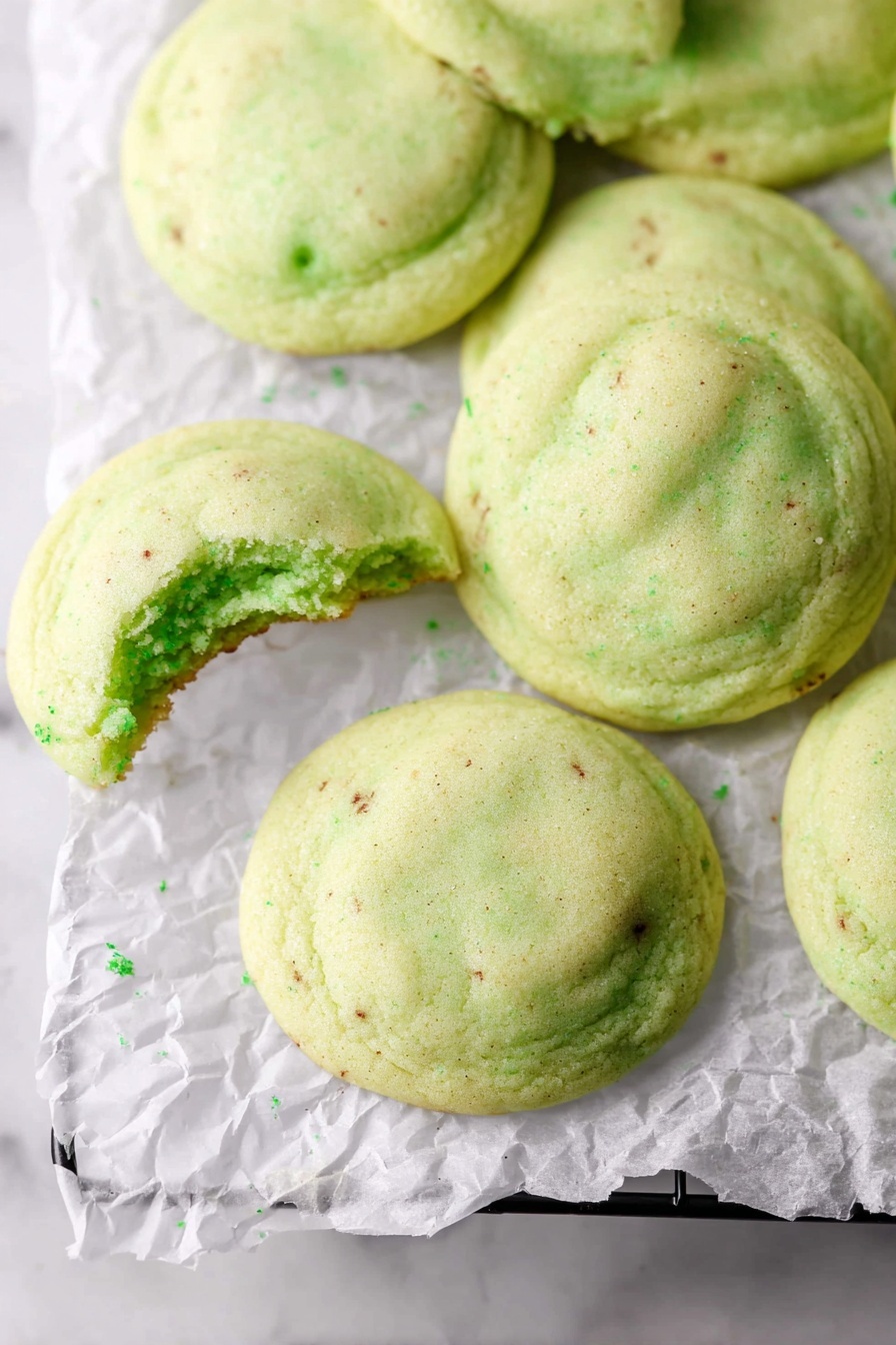 Pistachio Pudding Cookies, pistachio cookies, green pudding cookies, soft pistachio cookies, holiday cookie recipes - A group of soft, light green cookies with a smooth, slightly puffy texture are spread out on crinkled white parchment paper placed on a black wire rack. One cookie in the lower left corner has a bite taken out, showing a soft inside that matches the outer color. The cookies have small darker green spots scattered lightly on their surface. The whole setup is on a white marbled background. photo taken with an iphone --ar 2:3 --v 7